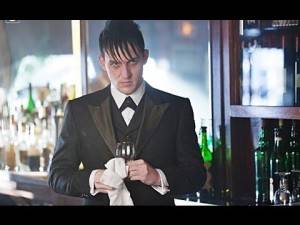 Scowling man in a tuxedo stands behind a bar, wiping a glass