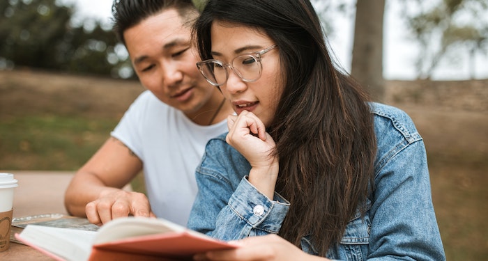 An Asian man and woman are both leaning over a book, reading intensely