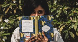 Indian woman holds a book to her face, obscuring her nose and mouth, and also showing her silver fingernails. She's standing in front of a a flowery bush