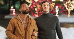 a still from Single All the Way, showing a Black and white man arm in arm with Christmas lights in the background