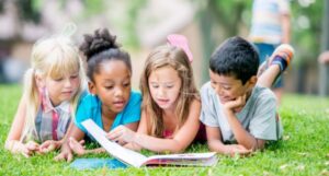 three girls and one boy of varying skin tones lie on the grass reading from the same book