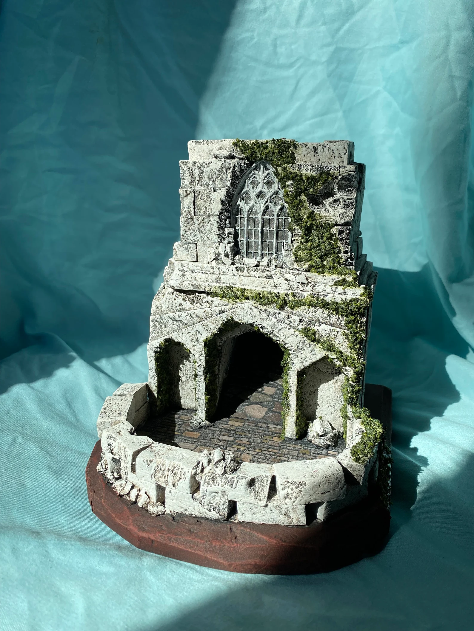 A photo of a gray Cathedral Dice Tower with a stained glass window and moss details.
