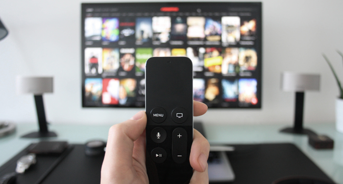 a hand holding a remote with a TV on a streaming service menu out of focus in the background