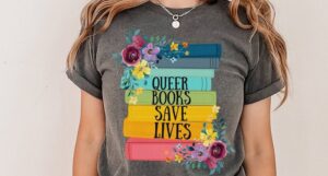 A photo of someone wearing a tee shirt with an illustration of a stack of rainbow colored book spines, florals, and the words "Queer books save lives."
