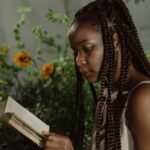 dark brown-skinned woman with braids reads a boot in a lush green garden