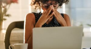 a brown-skinned woman with her hands held up to her face in a stressed expression working over a laptop