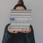 a fair-skinned person with their hair in a bun holding a stack of books that is obscuring their face