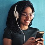 light brown-skinned Black woman with thick curly hair is wearing headphones, looking at her phone, and smiling
