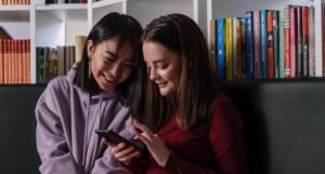a fair-skinned white woman and Asian woman looking and smiling at a smartphone while sitting in a bookstore