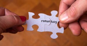 hands holding two connected puzzle pieces. one contains the letters r-o-m-a-n and the other t-a-s-y, combining to spell romantasy