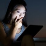 woman holding her hand up to her face in shock while reading from a tablet in the dark