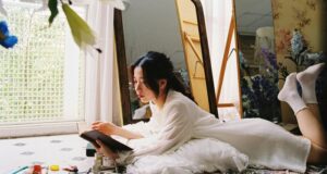 fair-skinned Asian woman lying on her stomach and reading a book in front of a floor-length mirror