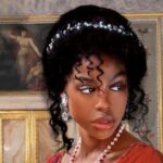 closeup of the cover of Aphrodite and the Duke by J.J. McAvoy, showing a Black woman in a red dress from the neck up. Her dark hair is piled high and held back with a band of jewels. She is wearing large diamond teardrop earrimgs and a string of pearls around her neck