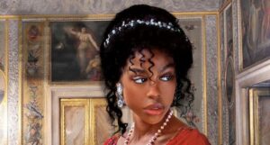closeup of the cover of Aphrodite and the Duke by J.J. McAvoy, showing a Black woman in a red dress from the neck up. Her dark hair is piled high and held back with a band of jewels. She is wearing large diamond teardrop earrimgs and a string of pearls around her neck
