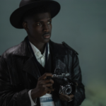 photo of a Black man holding an analog camera and wearing a press pass against a foggy background