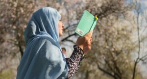 fair-skinned woman in hijab reading a book; there are blossoming trees in the background