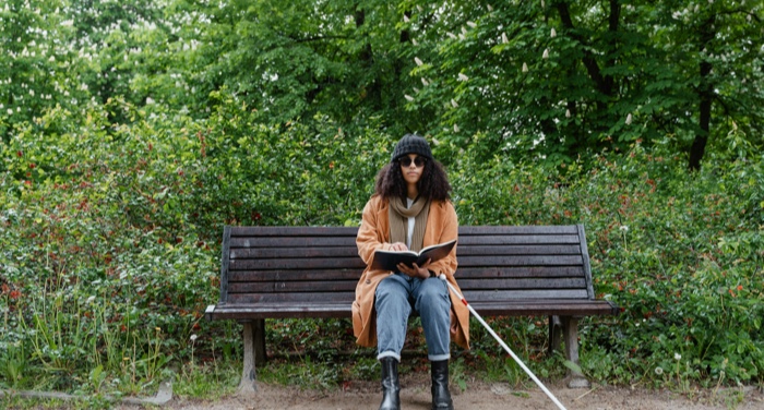 a tan-skinned Black woman sitting on a park bench and reading; she has a white cane used by visually impaired people