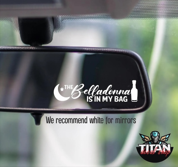 white vinyl decal on a rear view mirror. The decal is a half moon and a bottle of wine on either side of text that reads "the belladonna is in my bag"