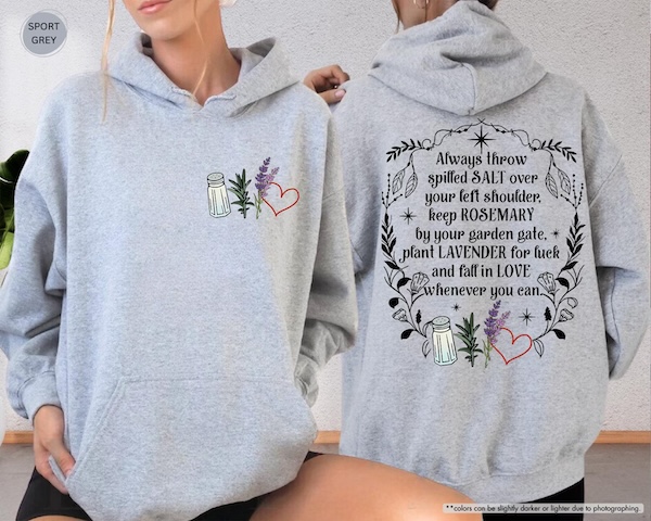 a light grey hooded sweatshirt. On the front are graphics of a salt shaker, a sprig of rosemary, a sprig of lavender, and a heart. On. the back is a quote from Practical Magic bordered by flowers and vines