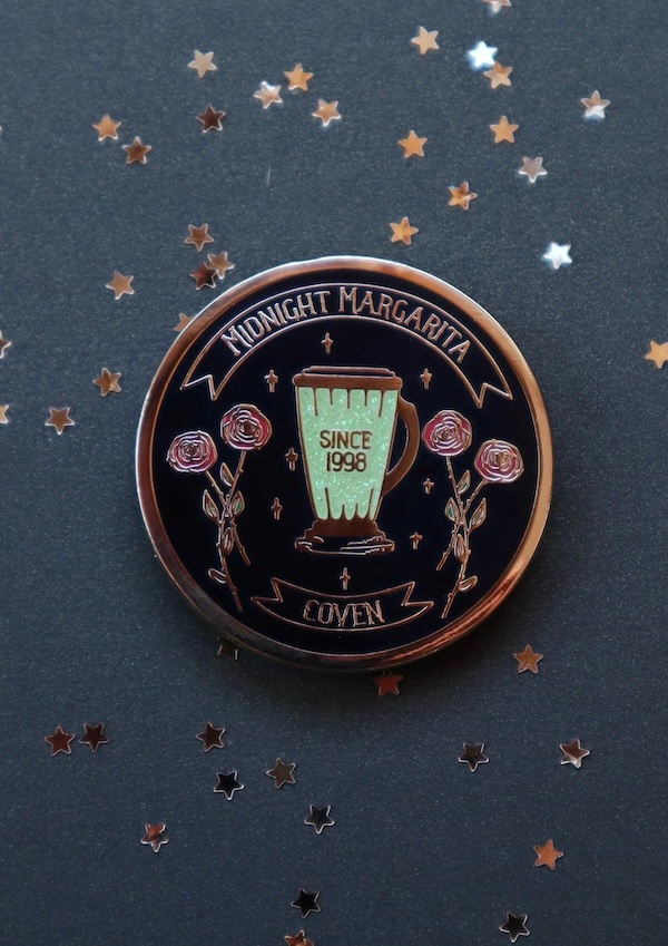 a black and brass enamel pin. In the center of the pin are a blender containing green liquid and two small bouquets of roses. Text bordering the design reads Midnight Margarita Coven