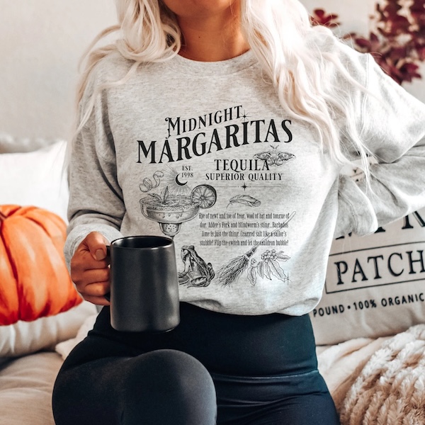 a light brown crewneck sweater with text and graphics in black ink. The text reads "Midnight Margaritas, Tequila, Superior Quality" and the graphics are of a margarita glass, a feather, a broom, a frog, and a belladonna plant