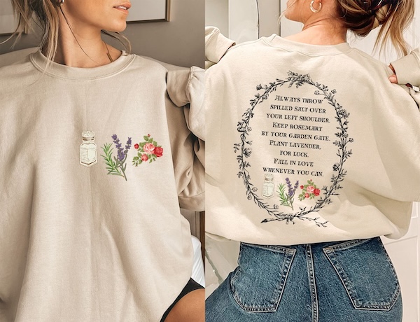 a sand-colored crewneck sweatshirt. On the front are graphics of a salt shaker, a sprig of rosemary, a sprig of lavender, and a heart. On. the back is a quote from Practical Magic bordered by flowers and vines