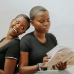 two brown-skinned Black women with short haair sitting back to back and reading books