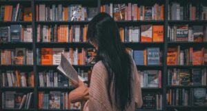 asian person reading a book in front of a filled bookshelf in a book store