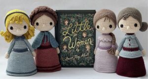 Four little crocheted aigurumi figures wearing historical dresses in the style of the American Civil War and designed to look like Amy, Meg, Beth, and Jo from Little Women. The figures are set around a beautiful green edition of the book, The Little Women.