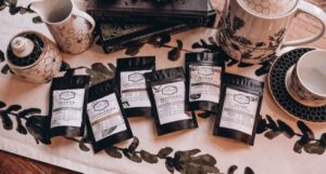 six black resealable pouches of loose leaf teas with white labels for Lord of the Rings themed tea. The blends are Gondor, Lothlorien, Misty Mountain, Rivendell, Rohan, and Shire, and the bags are on a table with a stack of Lord of the Rings books and assorted ceramics