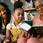 three generations of Black women (and one girl) reading a book on the couch