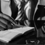 black and white photo of a person in a tie and trenchcoat writing in a notebook at a desk