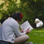a fair-skinned man reading a book outside on the lawn with swans in the background