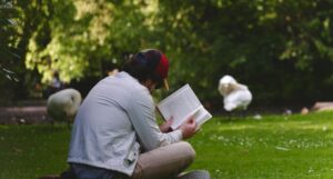 a fair-skinned man reading a book outside on the lawn with swans in the background