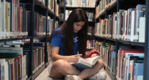fair-skinned teen with long dark hair reading while sitting on the floor in a library