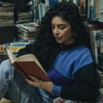 a light/medium skinned woman is sitting on the floor and reading in a used bookstore