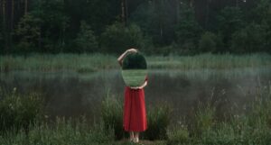 fair-skinned person in a red dress holds up an oval mirror in front of their face and torso while standing outside