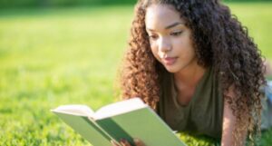 Image of a Black teenager reading a green book outside.