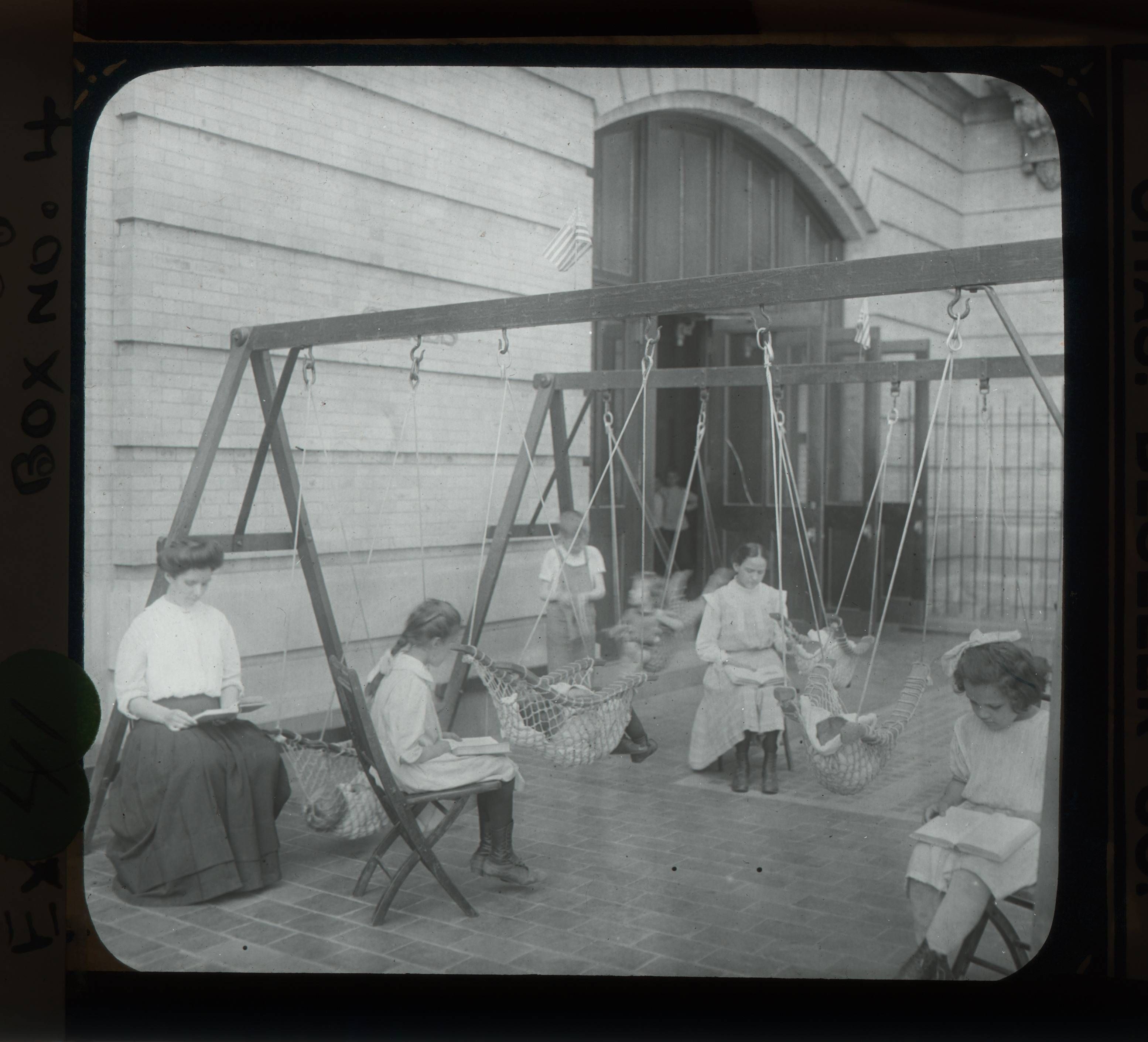 "Women and girls reading near swingset where younger children are suspended in hammock like swings, July 1910" - Lewis Wickes Hine