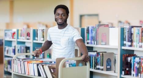 Portrait of young male librarian with trolley of books in library