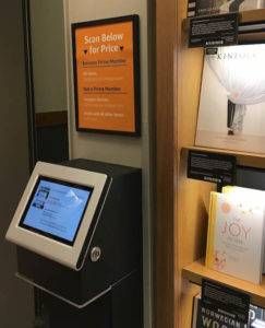 An electronic, touch screen kiosk beside a bookself. A sign above it offers instructions