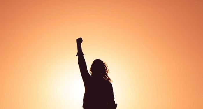female figure raising fist triumphantly against orange sunrise-like backdrop