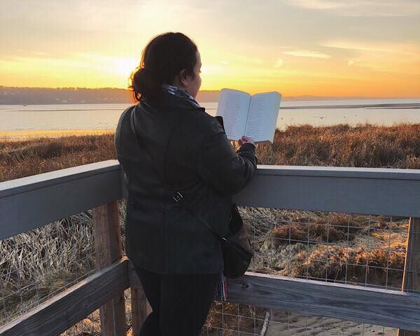 A woman reading, facing away, in front of a waterfront sunset