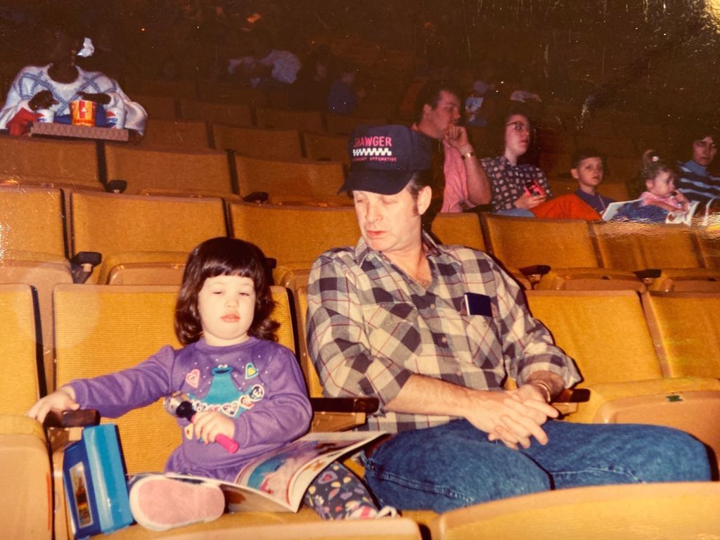 The author as a toddler, in a purple Cookie Monster outfit, sits next to her father waiting for Sesame Street Live. 