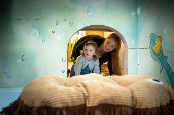 White woman and white girl poke their heads through a small opening into a room with bubbles and sea creatures on the walls