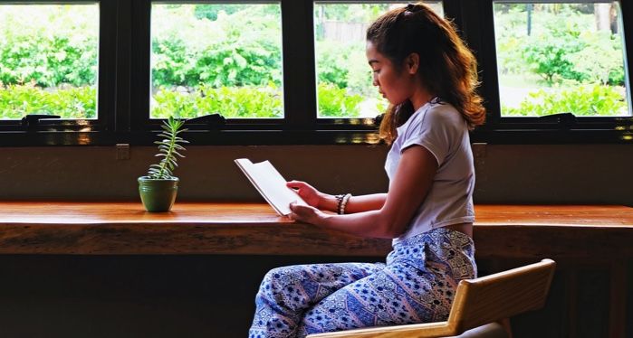 young woman reading a book at a counter in front of windows revealing greenery