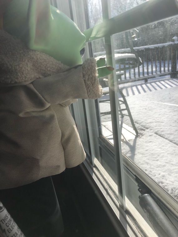 Grogu plush toy looks out of window at snow covered deck dreaming of reading children's books about snow; photo by author