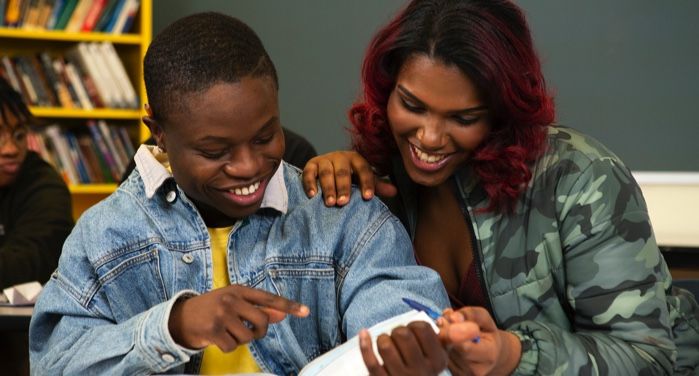 a brown-skinned Black transfeminine student and brown-skinned Black masculine student laughing together while reading