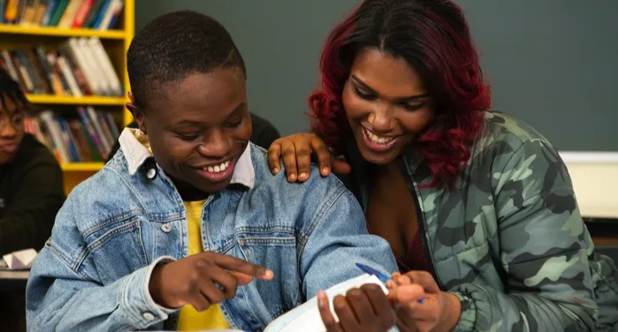 a brown-skinned Black transfeminine student and brown-skinned Black masculine student laughing together while reading