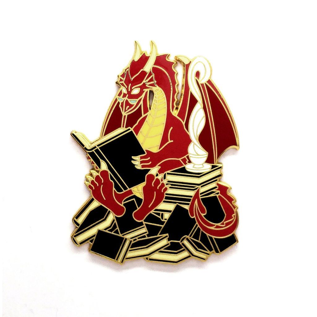 An enamel pin of a dragon sitting on a stack of books, reading, with a steaming mug beside it